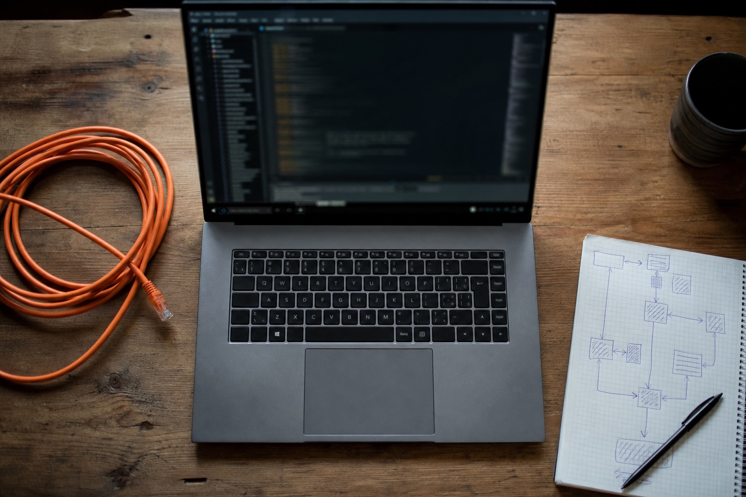 Plano cenital de un portátil sobre mesa de madera con terminal abierta, cable de red desenrollado y libreta con diagramas dibujados a mano